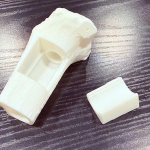 May include: A white 3D printed object resembling a hollowed-out tree branch with a textured bark-like exterior. The main piece features a cylindrical opening and a rectangular cutout, accompanied by a separate, smaller curved white piece.