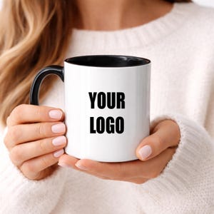 May include: A white ceramic mug with a black interior and a black handle. The exterior of the mug features the black text YOUR LOGO printed in a bold, sans-serif font. The mug has a cylindrical shape with a smooth, glossy finish throughout.