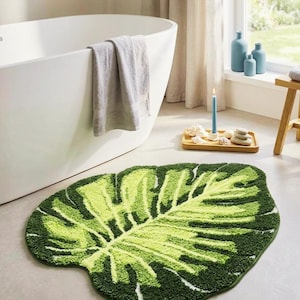 May include: A tufted bath mat shaped like a large monstera leaf, featuring a gradient of vibrant lime green and deep forest green colors with detailed cut-out sections and a textured, plush pile surface that mimics the natural veins of a leaf.