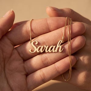 May include: A gold-colored name necklace featuring the name Sarah in a cursive script font. The pendant is attached to a delicate box chain, creating a simple and elegant piece of jewelry that rests horizontally across the center of the design.