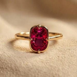 May include: A gold ring featuring a central rectangular cushion-cut vibrant red gemstone held in a four-prong setting. The band is thin and polished, showcasing a smooth metallic finish that complements the deep red color of the stone.