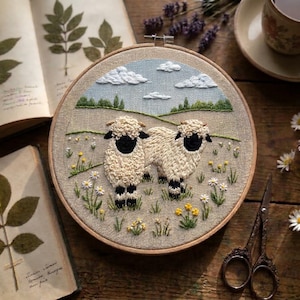 May include: A circular embroidery hoop featuring a hand-stitched landscape of two fluffy sheep with black faces and legs standing in a field of white and yellow wildflowers under a light blue sky with white clouds, set against a beige fabric background.