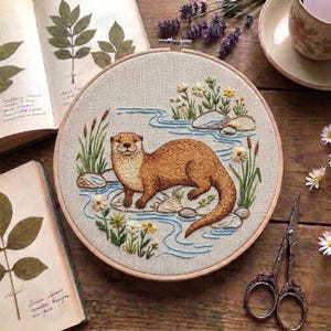 Puede incluir: Un bastidor de bordado circular que presenta una nutria marrón cosida a mano descansando sobre una roca en un arroyo azul. La escena incluye juncos verdes, pequeñas flores blancas y amarillas y piedras grises sobre una tela clara.