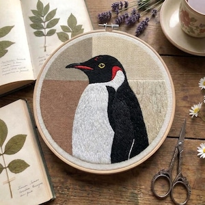 Könnte beinhalten: Ein runder Stickrahmen mit einem detaillierten, mit Faden gestickten Pinguin mit schwarzem, weißem und rotem Gefieder. Der Hintergrund ist in vier geometrische Quadranten aus beigem, hellbraunem und cremefarbenem Stoff unterteilt.