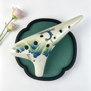 May include: A cream-colored ceramic ocarina featuring a hand-painted blue and green mountain landscape design. The instrument has multiple finger holes, the letters AC embossed on the front, and delicate black ink calligraphy near the base.