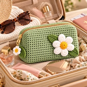 Può includere: Una pochette all'uncinetto color verde salvia con una grande applicazione a forma di margherita bianca con centro giallo e due foglie verdi. La pochette presenta una chiusura a zip dorata con un piccolo ciondolo a margherita abbinato.