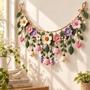 May include: A wall hanging featuring a crocheted garland of flowers, leaves, and hearts in shades of pink, purple, white, and green. The arrangement is suspended from a thick jute rope with wooden beads and includes various floral shapes.
