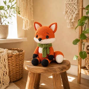 May include: A crocheted fox stuffed animal with bright orange fur, a white chest and face patch, black paws, and a white-tipped tail. The fox wears a textured olive green scarf around its neck and features round black eyes and a small black nose.