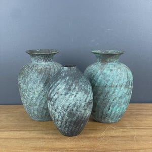 May include: Three vases with a weathered, oxidised teal and dark grey patina. Two vases feature a wide, flared rim and a rounded body, while the central vase has a narrower, tapered neck and a more elongated, oval-shaped body structure.