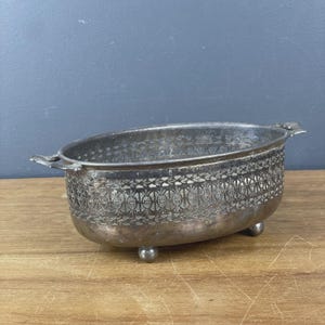 May include: An oval silver-toned metal bowl featuring an intricate openwork lattice pattern along the sides. The bowl has two small handles on opposite ends and is supported by four small, rounded feet, showcasing a weathered metallic finish.