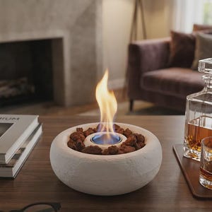 May include: A round, white, textured stone tabletop fire pit featuring a central burner surrounded by dark brown volcanic rocks. A bright orange and blue flame rises from the centre of the circular basin, which sits on a flat wooden surface.