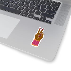 Könnte beinhalten: Ein gestanzter Aufkleber, der eine braun getönte Hand zeigt, die ein Friedenszeichen macht. Die Hand ist mit dunkelrotem Nagellack und einem breiten, texturierten pinkfarbenen Armband mit goldfarbenen Akzenten verziert, auf einer silbernen Laptop-Oberfläche.
