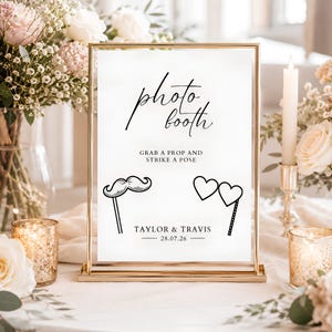 Può includere: Un cartello rettangolare con cornice dorata e carta bianca con testo nero che recita photo booth, GRAB A PROP AND STRIKE A POSE, TAYLOR & TRAVIS e 28.07.26. Il cartello include disegni a linee nere di un baffo e occhiali a forma di cuore.