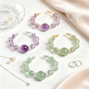 Könnte beinhalten: Vier offene Armreifen mit runden Edelsteinperlen in Violett und Grün, die mit Metalldraht umwickelt sind. Zwei Armreifen enthalten violette Amethyst-artige Perlen, zwei enthalten grüne Aventurin-artige Perlen. Zwei kleine Ringe sind enthalten.