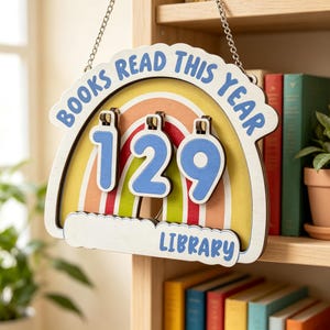 May include: A wooden wall hanging sign shaped like a rainbow with the text "BOOKS READ THIS YEAR" and "LIBRARY" in blue. It features three hanging blue number tiles displaying "129" suspended by a silver chain, designed for tracking book counts.