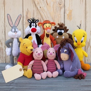 Puede incluir: Una colección de nueve juguetes de ganchillo hechos a mano que incluyen a Bugs Bunny, el gato Silvestre, Tigger, el Demonio de Tasmania, Piolín, Winnie the Pooh, dos figuras de Piglet y Igor, junto a una etiqueta de papel beige rectangular.