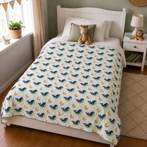 Puede incluir: Una colcha de cama blanca con un patrón repetitivo de ballenas azul oscuro y lunas crecientes naranjas. La colcha cubre una estructura de cama blanca, decorada con una almohada blanca, una almohada verde y un juguete de jirafa de peluche.