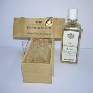 Puede incluir: Una botella de vidrio rectangular de Echt Kölnisch Wasser Farina Dem zu Mailand con tapa blanca, junto a una caja de madera abierta que contiene envoltorio de plástico transparente. La etiqueta muestra un escudo y el texto Echt Kölnisch Wasser.