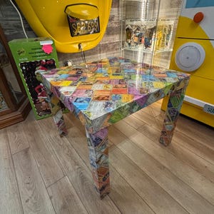May include: A square side table with a glossy finish, featuring a surface and four legs completely covered in a collage of colorful trading cards. The cards display various illustrations, text, and borders in shades of yellow, blue, pink, and green.