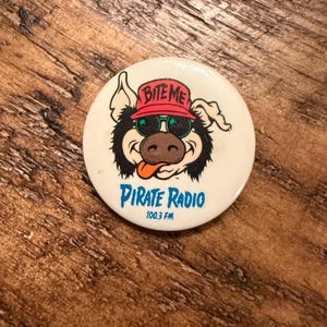 Puede incluir: Un botón redondo de color blanco con un cerdo de dibujos animados que lleva una gorra roja con el texto BITE ME, gafas de sol verdes y un hocico marrón. Debajo del cerdo, el texto dice PIRATE RADIO 100.3 FM en letras de color azul.