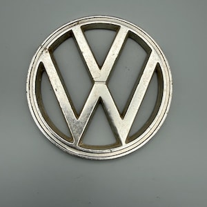 Może przedstawiać: Okrągły, srebrny emblemat Volkswagena z kultowymi literami V i W wyśrodkowanymi wewnątrz pierścienia. Metalowa powierzchnia wykazuje ślady zużycia, w tym lekkie zadrapania i otarcia, z matowym wykończeniem na całym logo.