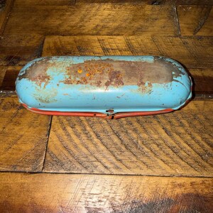 Könnte beinhalten: Ein längliches Brillenetui aus Metall im Vintage-Stil mit abgenutzter hellblauer Oberfläche, sichtbaren Rostflecken auf der Oberseite und einer kontrastierenden orange-roten Basis. Das Etui hat eine abgerundete, längliche Form mit Scharnier.