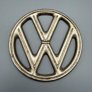 Puede incluir: Un emblema circular de metal plateado con el logotipo de Volkswagen, caracterizado por una V y una W estilizadas encerradas en un anillo. La superficie muestra signos de desgaste, incluyendo una ligera oxidación y textura de metal envejecido.
