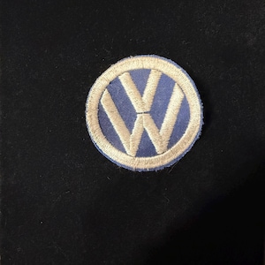 May include: A circular embroidered patch featuring the Volkswagen logo. The design consists of a blue background with a white V and W letter combination, enclosed within a thick, circular white stitched border on a solid black fabric surface.