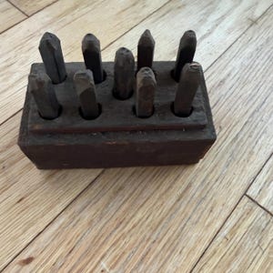 May include: A set of nine dark metal stamps arranged in a rectangular wooden block with pre-drilled holes. Each stamp features a distinct character or number on its top end, and the wooden holder has a weathered, dark brown finish.