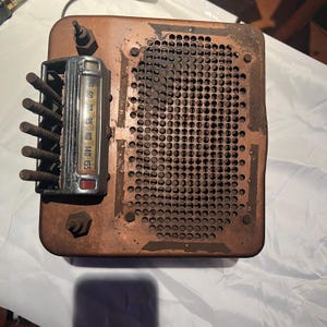 Op de afbeelding: Een vintage koperkleurige radio met een rechthoekige behuizing, een groot rond geperforeerd luidsprekerrooster en een verchroomd wijzerplaatpaneel. De wijzerplaat toont frequentiegetallen van 55 tot 155, met vier verticale knoppen aan de linkerzijde.