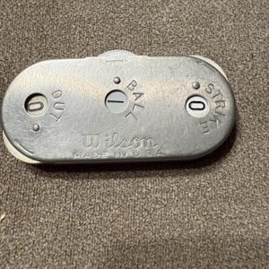Op de afbeelding: Een zilverkleurige metalen Wilson honkbal-scheidsrechterindicator met drie mechanische draaischijven voor het bijhouden van outs, ballen en strikes. Het apparaat bevat gegraveerde tekst met OUT, BALL, STRIKE, Wilson en MADE IN U.S.A. met cijfers.
