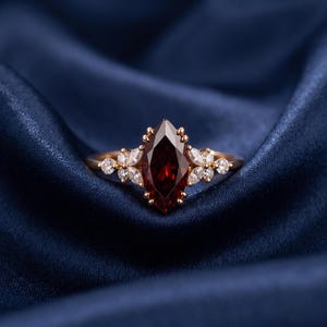 Op de afbeelding: Een gouden ring met een centrale dieprode edelsteen in marquise-slijpvorm, geflankeerd door clusters van kleine, heldere marquise- en rondgeslepen stenen aan weerszijden. De ringband is dun en metaalachtig met een gepolijste afwerking.