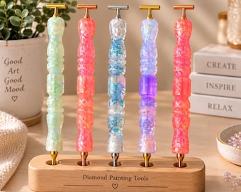 Könnte beinhalten: Fünf Diamond-Painting-Stifte mit glitzernden, durchscheinenden Harzgriffen in Hellgrün, Pink, Blau, Lila und Koralle, jeweils mit einem T-förmigen Metallaufsatz. Die Stifte stehen in einem rechteckigen Holzständer mit der Aufschrift Diamond Painting Tools.