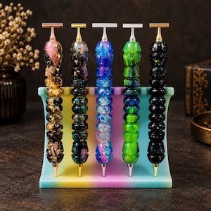 Op de afbeelding: Vijf pennen met gestructureerde, veelkleurige harsbehuizingen met metaalvlokken en glitters, elk met een T-vormige metalen clip. De pennen staan verticaal in een gebogen, regenboogkleurige standaard met vijf afzonderlijke sleuven voor de pennen.