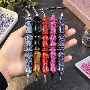 May include: Five diamond painting pens with ergonomic, multi-faceted glitter handles in blue, black, gold, red, and pink. Each tool features a metallic silver-toned angled tip holder at the top and a pointed metal nib at the bottom for placing gems.