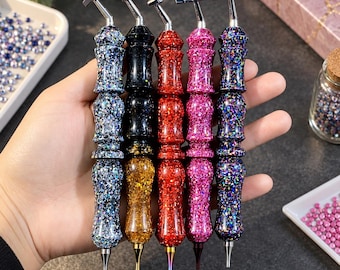 Könnte beinhalten: Fünf Diamond-Painting-Stifte mit ergonomischen, facettierten Glitzergriffen in Blau, Schwarz, Gold, Rot und Pink. Jedes Werkzeug verfügt über eine silberfarbene, abgewinkelte Halterung oben und eine feine Metallspitze zum Platzieren der Steinchen.