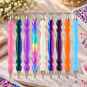 Può includere: Un set di nove penne per diamond painting disposte in fila, ognuna con un manico di colore unico tra cui rosa, verde, viola, arcobaleno, bianco, blu-rosa, nero, arancione e azzurro, tutte con punte e attacchi in metallo argentato.