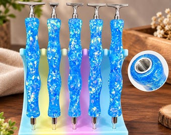 Könnte beinhalten: Fünf blaue Häkelnadeln aus Kunstharz mit irisierenden Flocken und silberfarbenen Metalloberteilen, aufrecht in einem pastellfarbenen Ständer. Ein kreisförmiger Ausschnitt zeigt das Ende einer Nadel mit einer Metallgewindeöffnung.