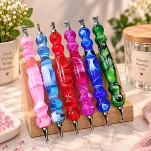 Op de afbeelding: Zes diamond painting pennen met ergonomische, gemarmerde acryl handvatten in roze, lichtblauw, rood, magenta, donkerblauw en groen. Elke pen heeft een zilverkleurige metalen punt en een metalen clip, rechtop geplaatst op een houten standaard.