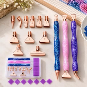 Op de afbeelding: Een diamond painting gereedschapsset met drie glitterende hars pennen in blauw, roze en paars. De set bevat diverse roségouden metalen opzetstukken genummerd 3 tot 10, een doorzichtige opbergdoos en verschillende kleine paarse wasblokjes.