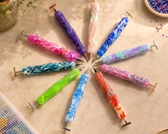 Könnte beinhalten: Eine Sammlung von zehn Diamond-Painting-Stiften, die kreisförmig angeordnet sind. Jeder Stift hat einen einzigartigen Schaft aus marmoriertem oder glitzerndem Harz in Farben wie Pink, Lila, Blau, Grün, Orange und Weiß mit Metallspitzen.
