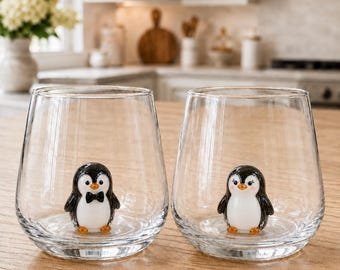Puede incluir: Dos vasos de cristal transparente, cada uno con una pequeña figura de pingüino de cristal en tres dimensiones fijada en la base interior. Un pingüino lleva una pajarita negra, mientras que el otro tiene pestañas delicadas y mejillas rosadas.