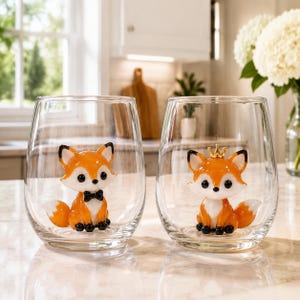 Peut inclure: Deux verres à vin sans pied transparents, contenant chacun une petite figurine de renard en trois dimensions. Le renard de gauche porte un nœud papillon noir, tandis que celui de droite porte une petite couronne dorée. Ils sont orange et blancs.