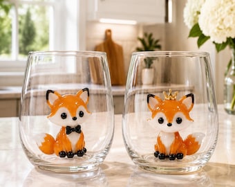 Peut inclure: Deux verres à vin sans pied transparents, contenant chacun une petite figurine de renard en trois dimensions. Le renard de gauche porte un nœud papillon noir, tandis que celui de droite porte une petite couronne dorée. Ils sont orange et blancs.
