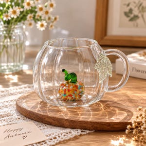 May include: A clear glass mug shaped like a pumpkin with vertical ridges and a handle. Inside the mug sits a small, colourful glass pumpkin figurine with a green stem. A textured, light green glass leaf is attached to the side of the mug handle.