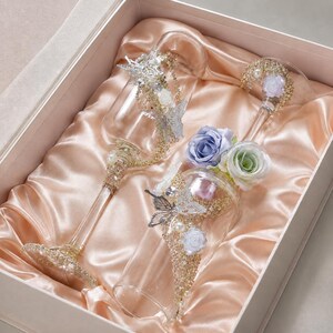 Peut inclure: Deux flûtes en verre transparent ornées d'accents papillons argentés, de paillettes dorées, de petites perles blanches et de roses artificielles bleues et vertes. Les verres sont présentés dans une boîte rectangulaire doublée de satin pêche.