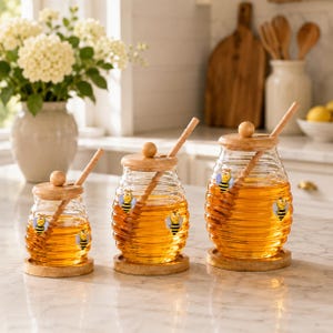 May include: Three clear glass honey jars of varying sizes, each featuring a ribbed beehive shape, a wooden lid with a knob, a wooden honey dipper, and a circular wooden base. Each jar is decorated with small, colorful bee illustrations.