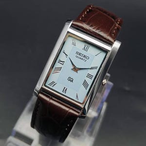 May include: A rectangular Seiko Quartz watch with a silver-tone case and a brown leather strap featuring a textured pattern. The watch face is light blue with a central wave-like design, black Roman numeral hour markers, and black hands.