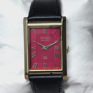 Könnte beinhalten: Eine rechteckige Seiko Quartz Armbanduhr mit einem leuchtend magentafarbenen Zifferblatt mit konzentrischem Texturmuster, goldenen römischen Ziffern als Stundenmarkierungen, goldenen Zeigern und einem polierten goldfarbenen Gehäuse mit schwarzem Lederarmband.