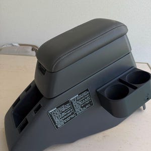Op de afbeelding: Een donkergrijze middenconsole voor in de auto met een gevoerd armsteundeksel, twee geïntegreerde bekerhouders aan de zijkant en een rechthoekig waarschuwingslabel met witte tekst. De unit bevat open opbergvakken en een kunststof afwerking.